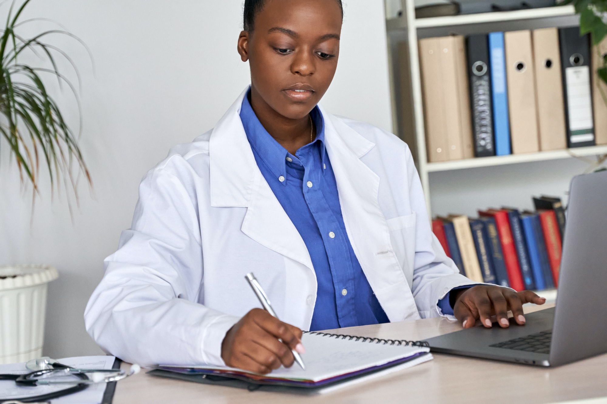 African american doctor writing medical record in notebook using laptop at desk.